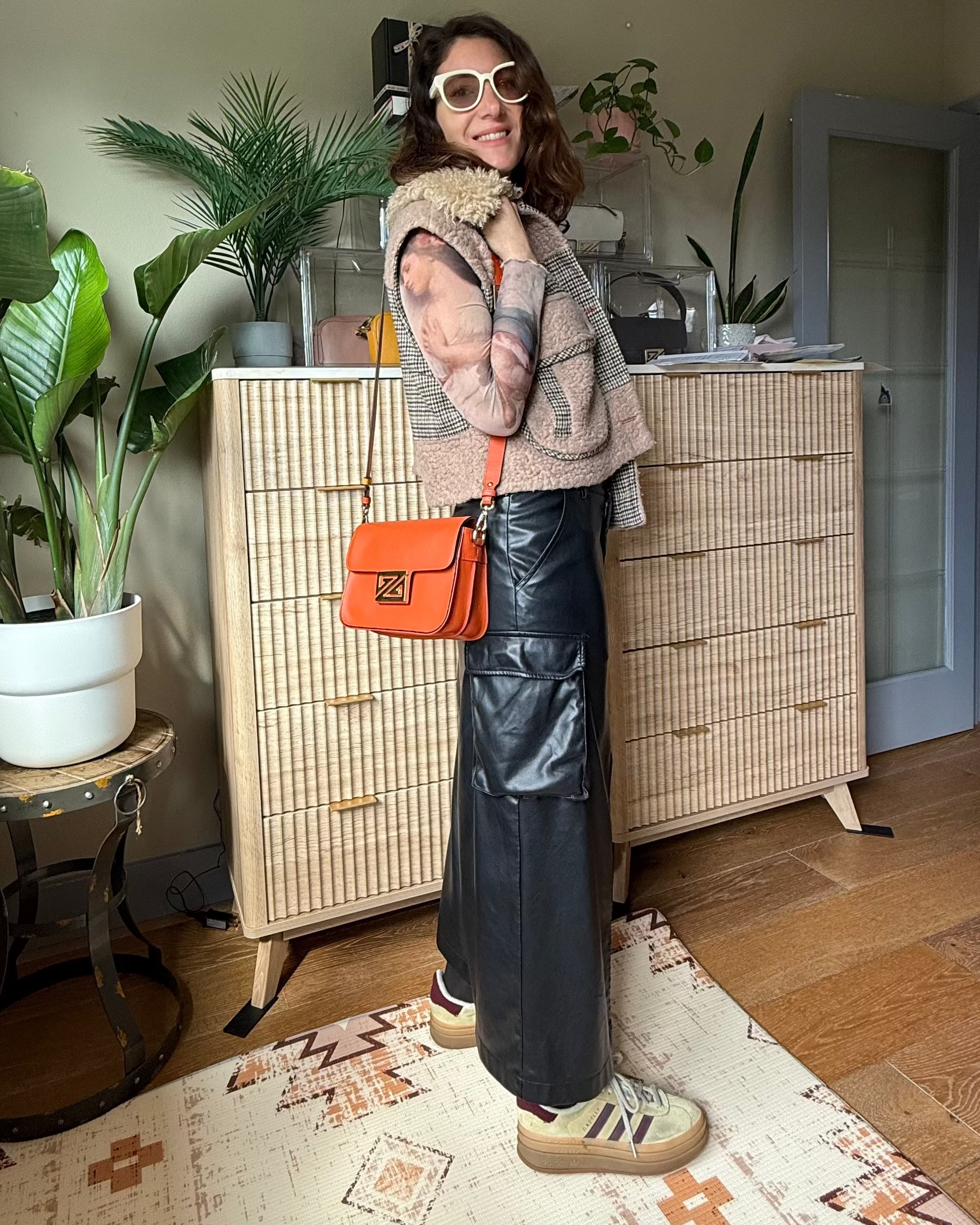 Woman standing in a room with plants and a dresser, wearing sunglasses and an orange bag.