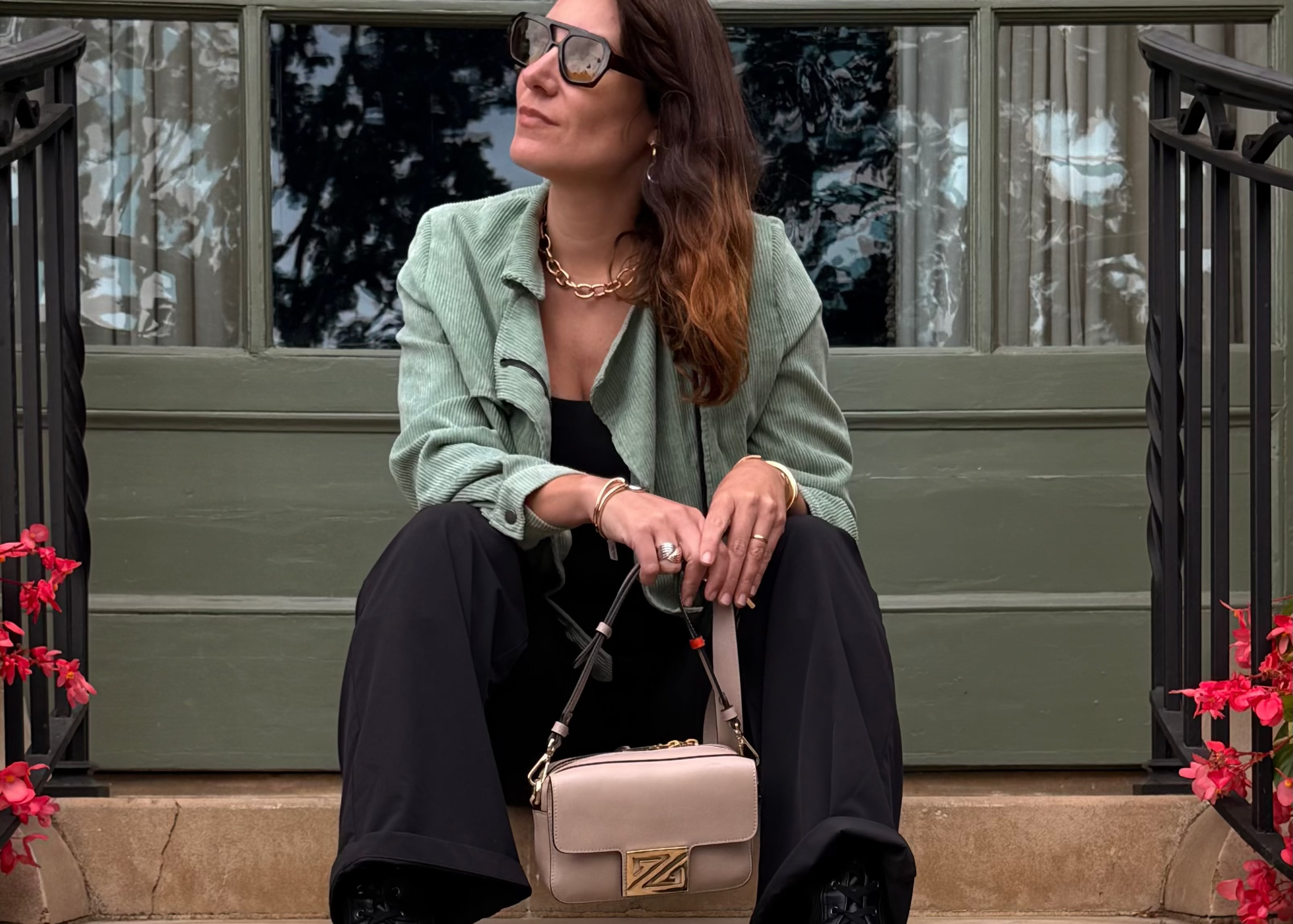 Woman sitting on steps wearing sunglasses and a green jacket with a pink handbag.