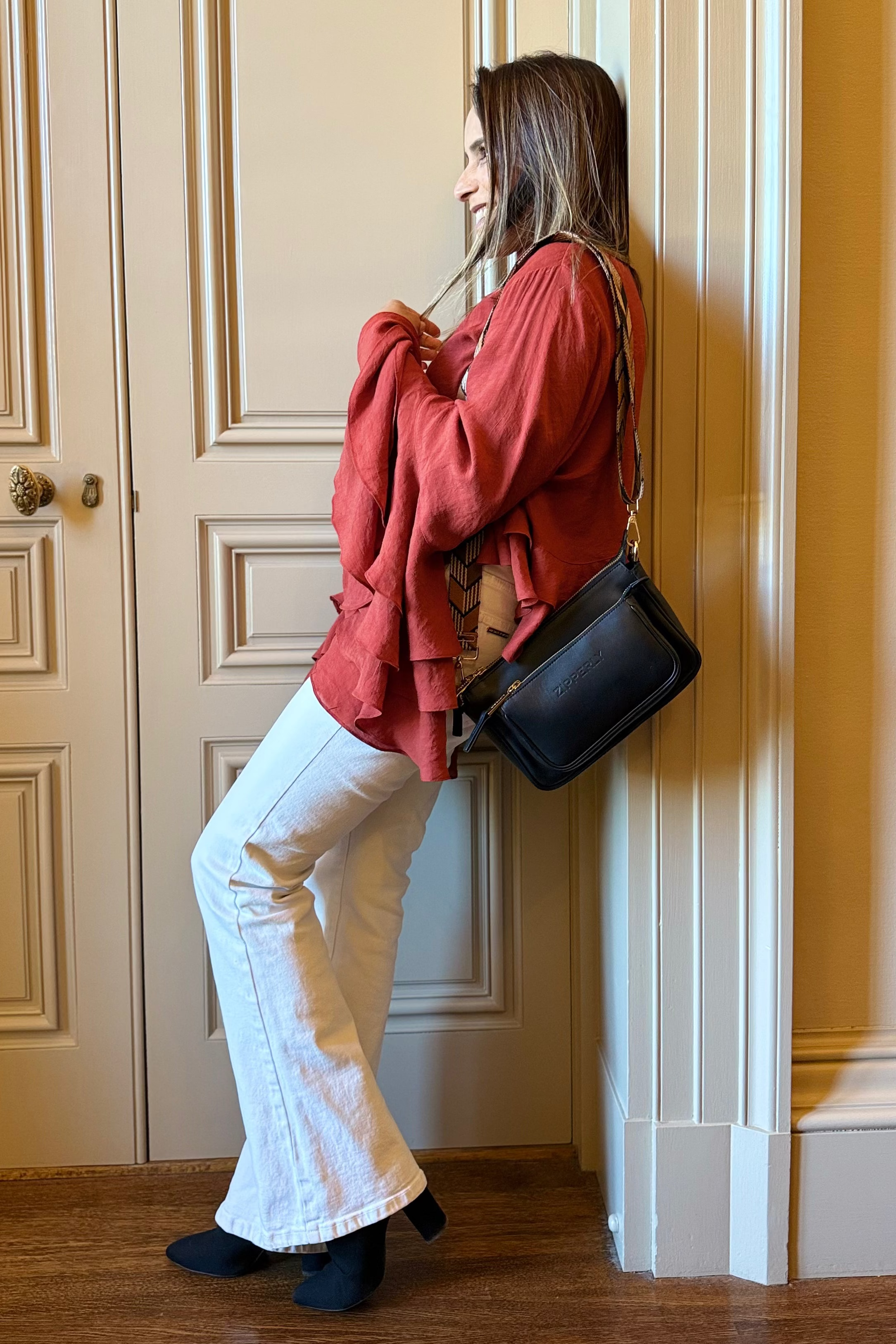 Woman in red sweater and white pants leaning against a wall with a black bag.