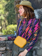 Woman wearing a colorful patterned top and yellow bag, leaning against a stone wall with greenery in the background.