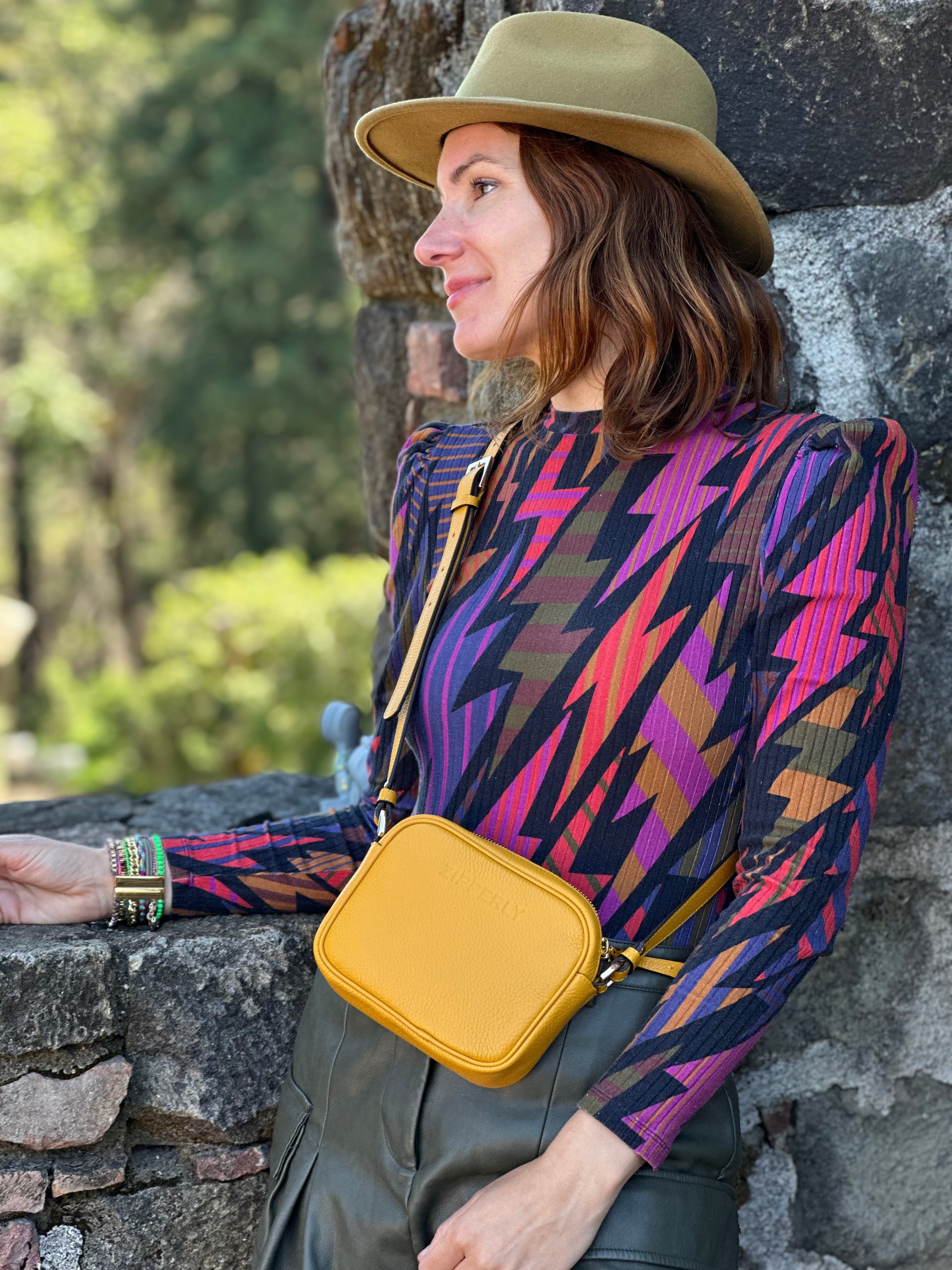 Woman wearing a colorful patterned top and yellow bag, leaning against a stone wall with greenery in the background.